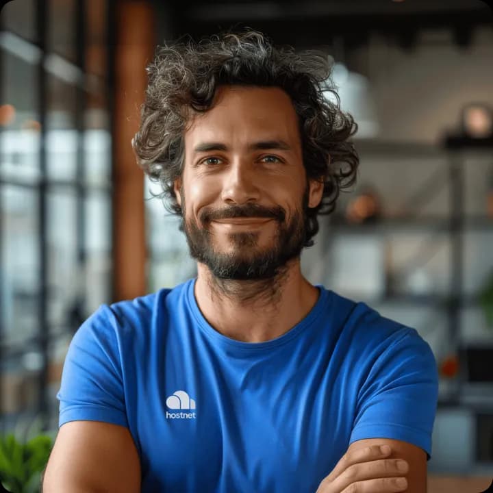 Man in een blauw T-shirt met het Hostnet-logo, glimlachend met de armen over elkaar in een kantoor.