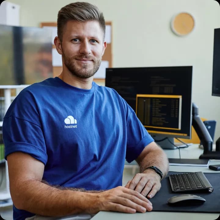 Glimlachende man in een blauw Hostnet-shirt aan een bureau met een toetsenbord en beeldschermen met code.